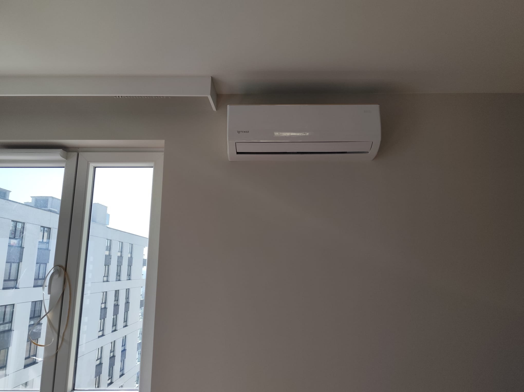 Biały klimatyzator Rotenso zamontowany na ścianie w jasnym pokoju, widok z okna na budynek mieszkalny. Minimalistyczny design, nowoczesne wnętrze.