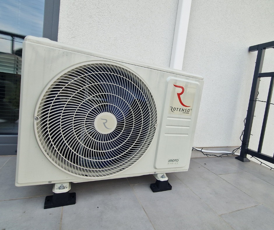 Biała jednostka zewnętrzna klimatyzacji Rotenso Imoto Series, zamontowana na balkonie, widoczna z boku, z elementami balustrady w tle. Lekko zabrudzona obudowa.