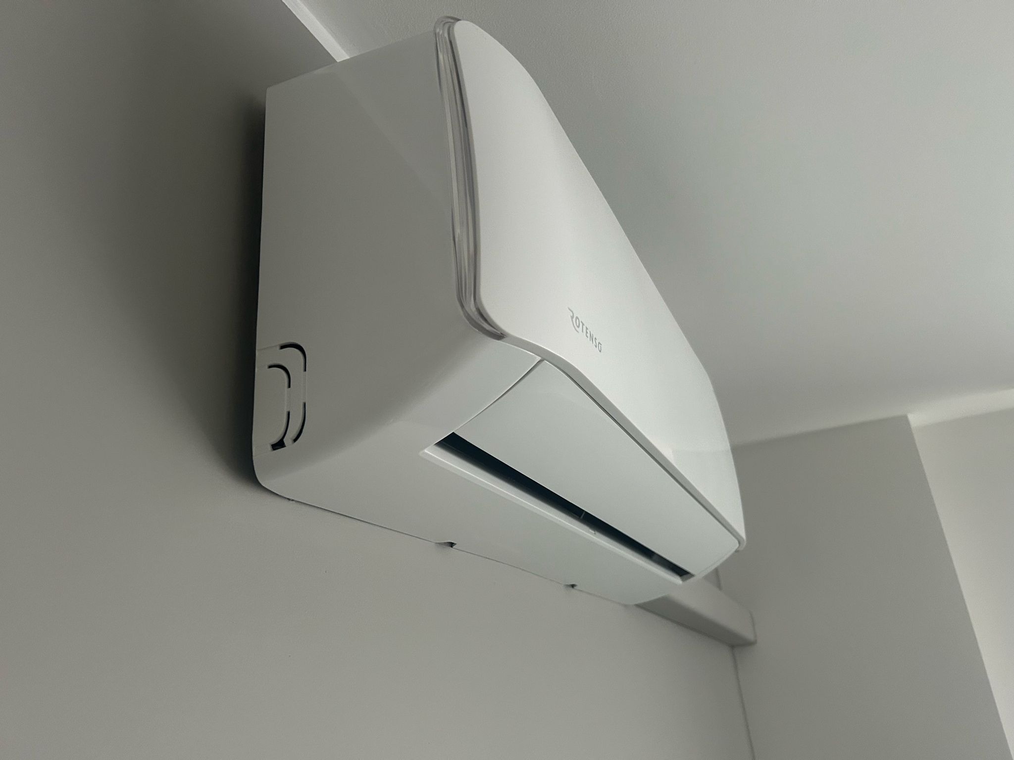 Biały klimatyzator Rotenso zamontowany na szarej ścianie, widok z dołu pod kątem. Minimalistyczny design, nowoczesne wnętrze.
