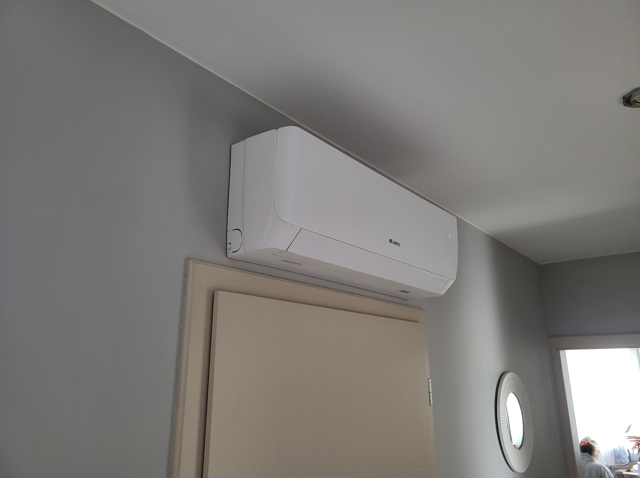 Biała klimatyzacja marki Gree zamontowana nad drzwiami w szarym pokoju. Widoczne okrągłe lustro i osoba w tle. Minimalistyczny design wnętrza.