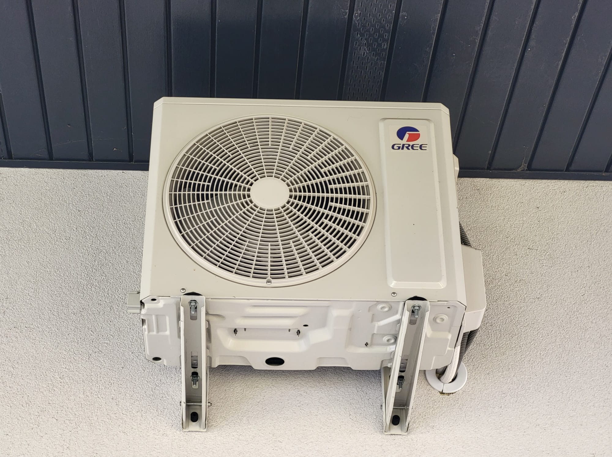 Zamontowany klimatyzator marki Gree na elewacji w Pińczycach. Widoczne wsporniki i przewody. Szara ściana i ciemne panele elewacyjne w tle. Ujęcie z dołu.