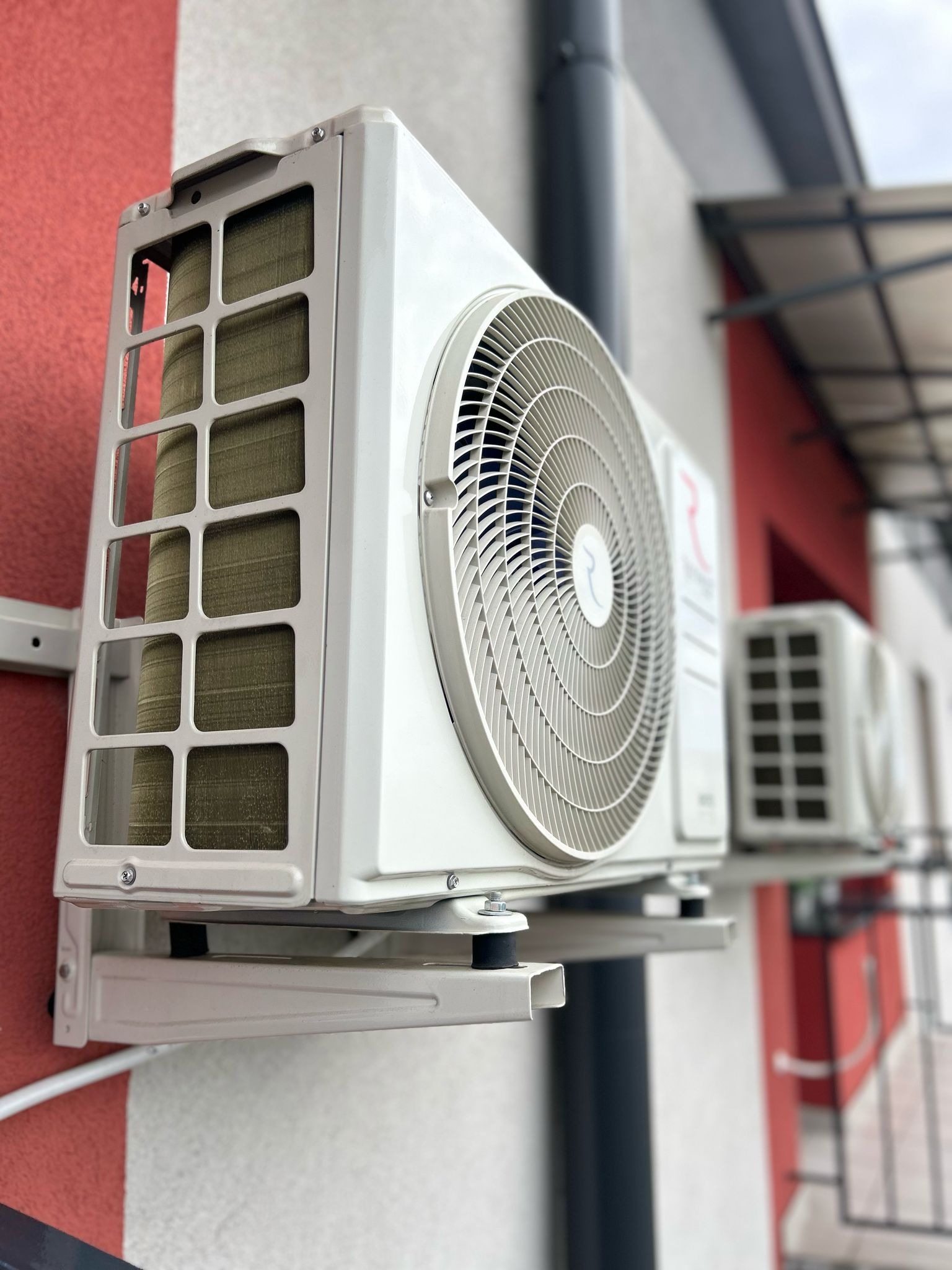 Zamontowana, biała jednostka zewnętrzna klimatyzacji na ścianie budynku z elementami czerwieni. Widoczna druga jednostka w tle, pod zadaszeniem.