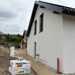 Ocieplenia - Biała elewacja domu jednorodzinnego w trakcie wykończenia, widoczne ocieplenie, tynk i elementy instalacji. Na pierwszym planie paczki styropianu.