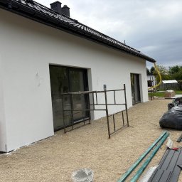 Ocieplenia - Biała elewacja domu jednorodzinnego po tynkowaniu, z widocznymi oknami, rusztowaniem i elementami wykończeniowymi na żwirowym podłożu. Widok z boku, pochmurne niebo.