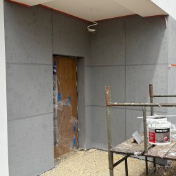 Ocieplenia - Elewacja budynku w trakcie tynkowania z efektem betonu, widoczne rusztowanie, wiadro z farbą 'art BETON' i narzędzia. Zabezpieczone oklejenie.