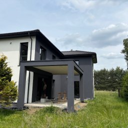 Ocieplenia - Dwukondygnacyjny dom z płaskim dachem i pergolą, malowany na biało i szaro. Widoczny mężczyzna malujący elewację. Trawa i drzewa w tle.