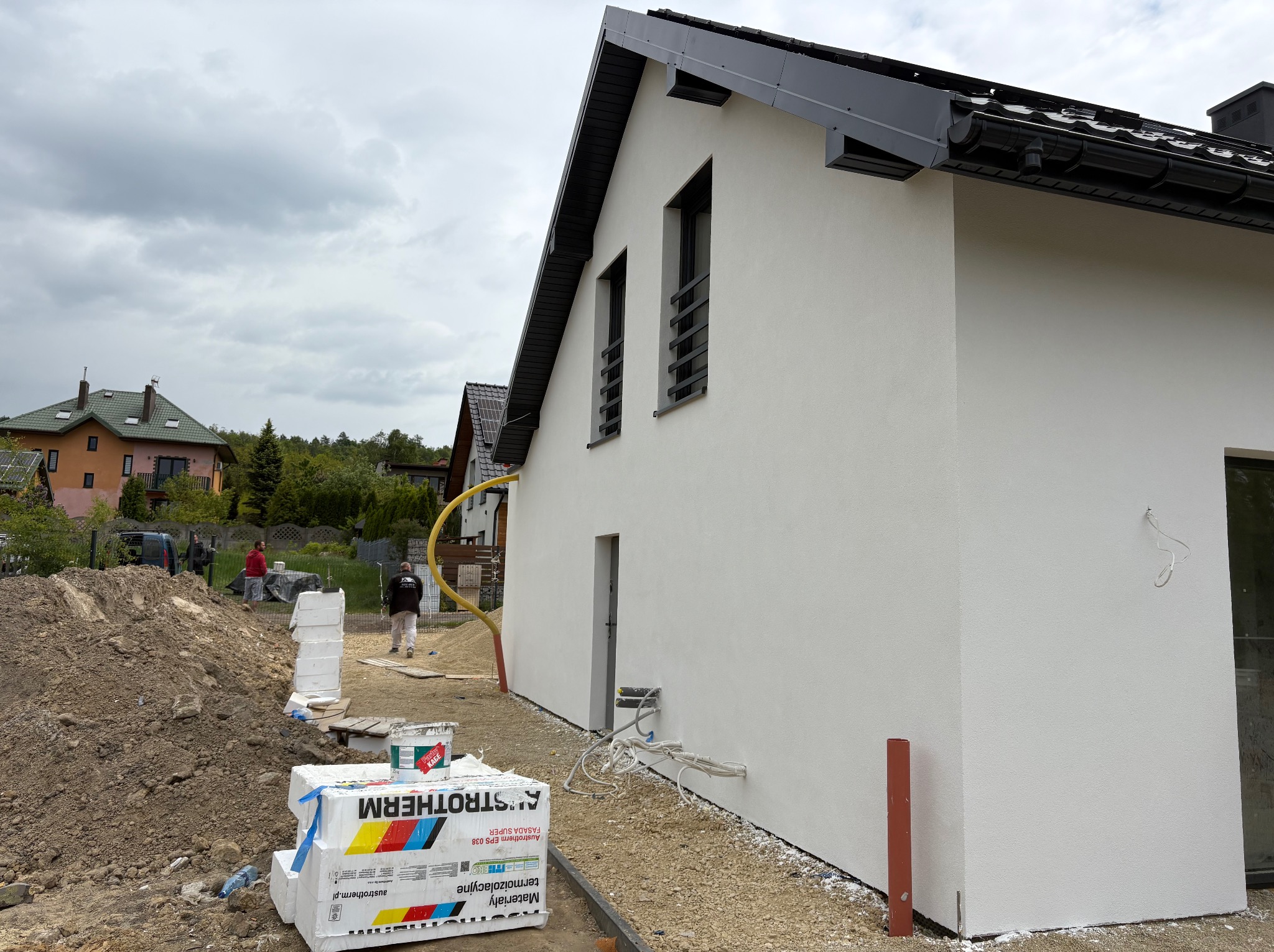 Biała elewacja domu jednorodzinnego w trakcie wykończenia, widoczne ocieplenie, tynk i elementy instalacji. Na pierwszym planie paczki styropianu.