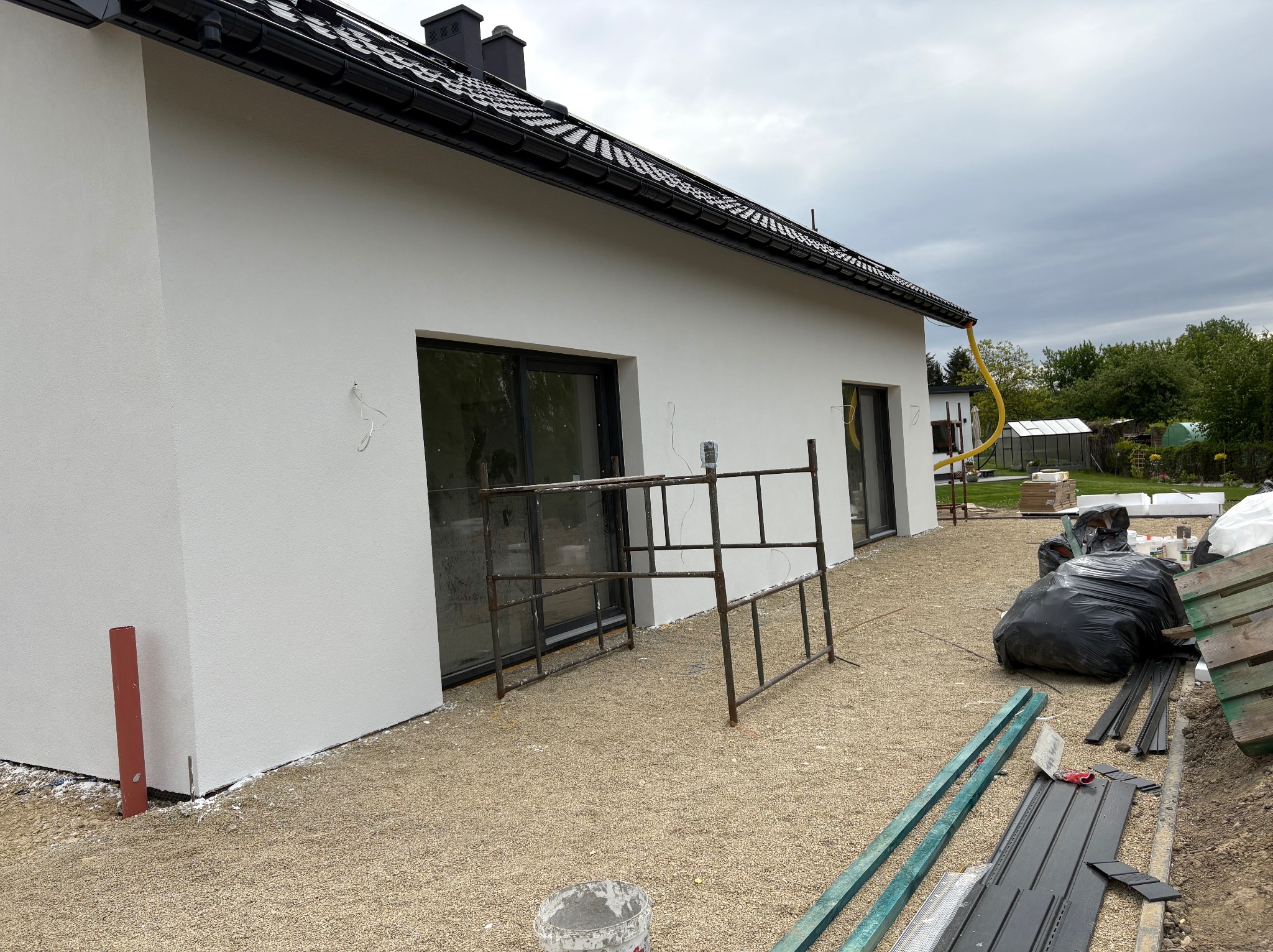 Biała elewacja domu jednorodzinnego po tynkowaniu, z widocznymi oknami, rusztowaniem i elementami wykończeniowymi na żwirowym podłożu. Widok z boku, pochmurne niebo.