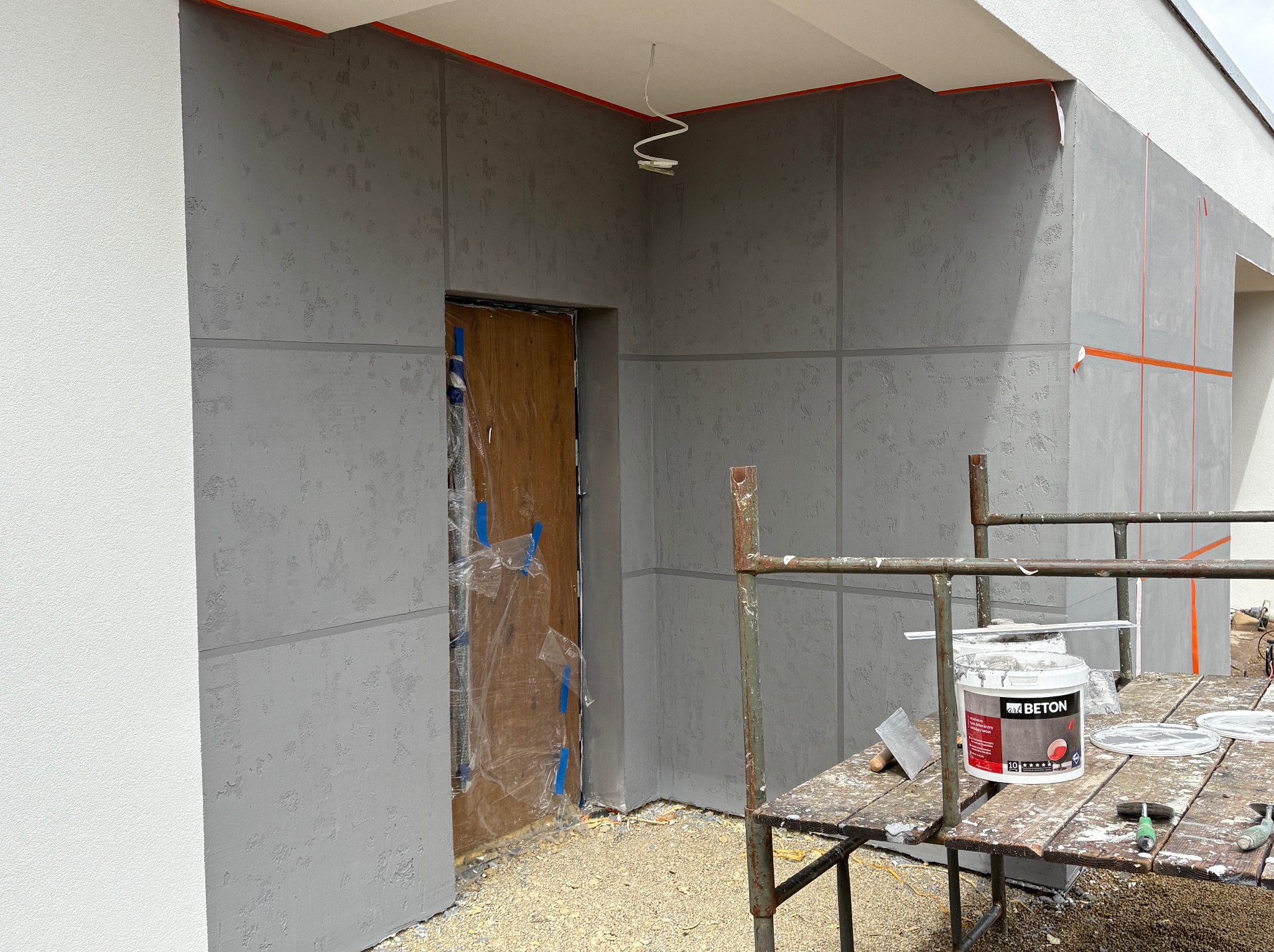 Elewacja budynku w trakcie tynkowania z efektem betonu, widoczne rusztowanie, wiadro z farbą 'art BETON' i narzędzia. Zabezpieczone oklejenie.