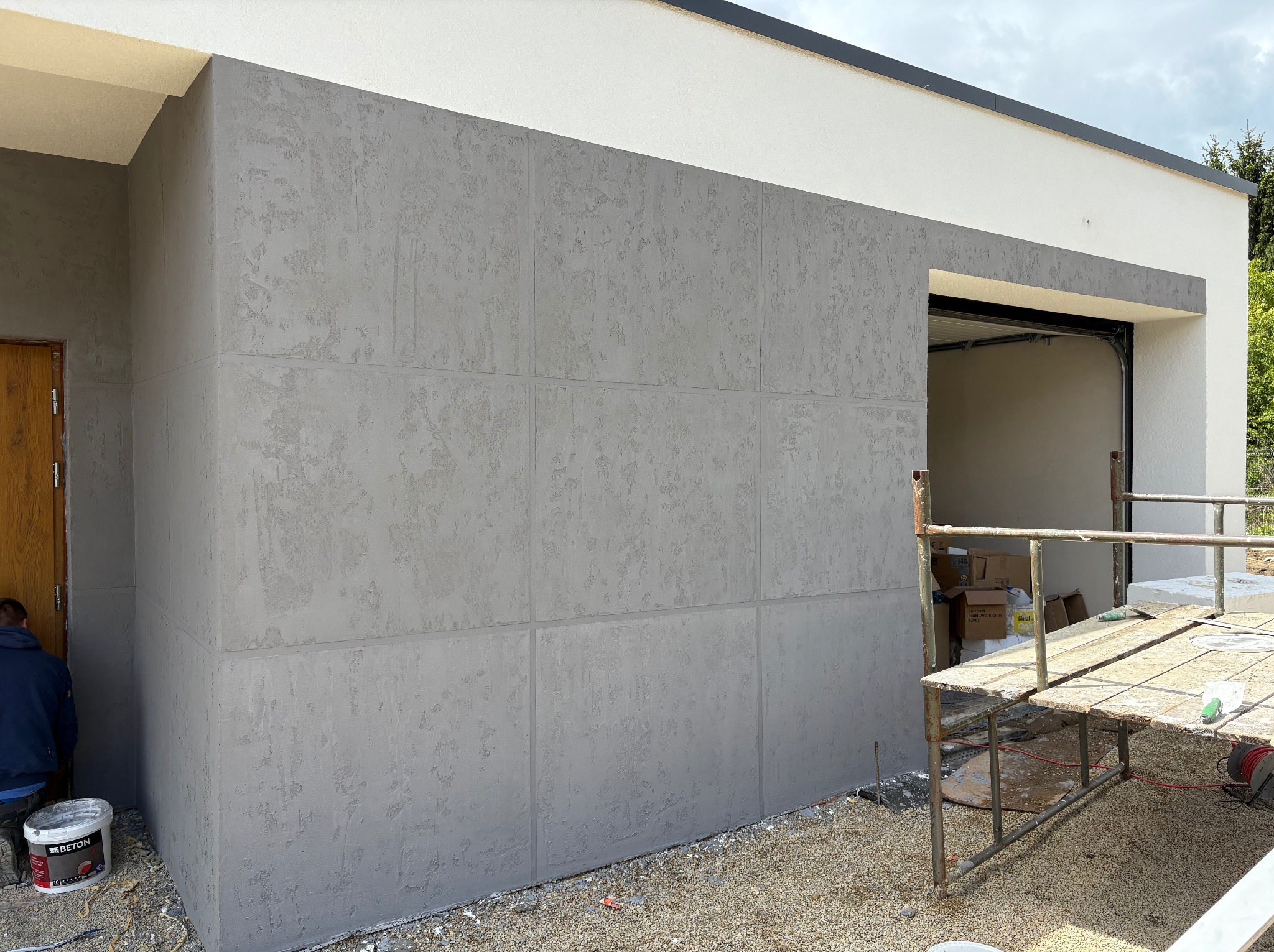 Szara elewacja budynku z efektem betonu architektonicznego, widoczny fragment rusztowania i pracownik przy wiadrze z farbą. Nowoczesny design.