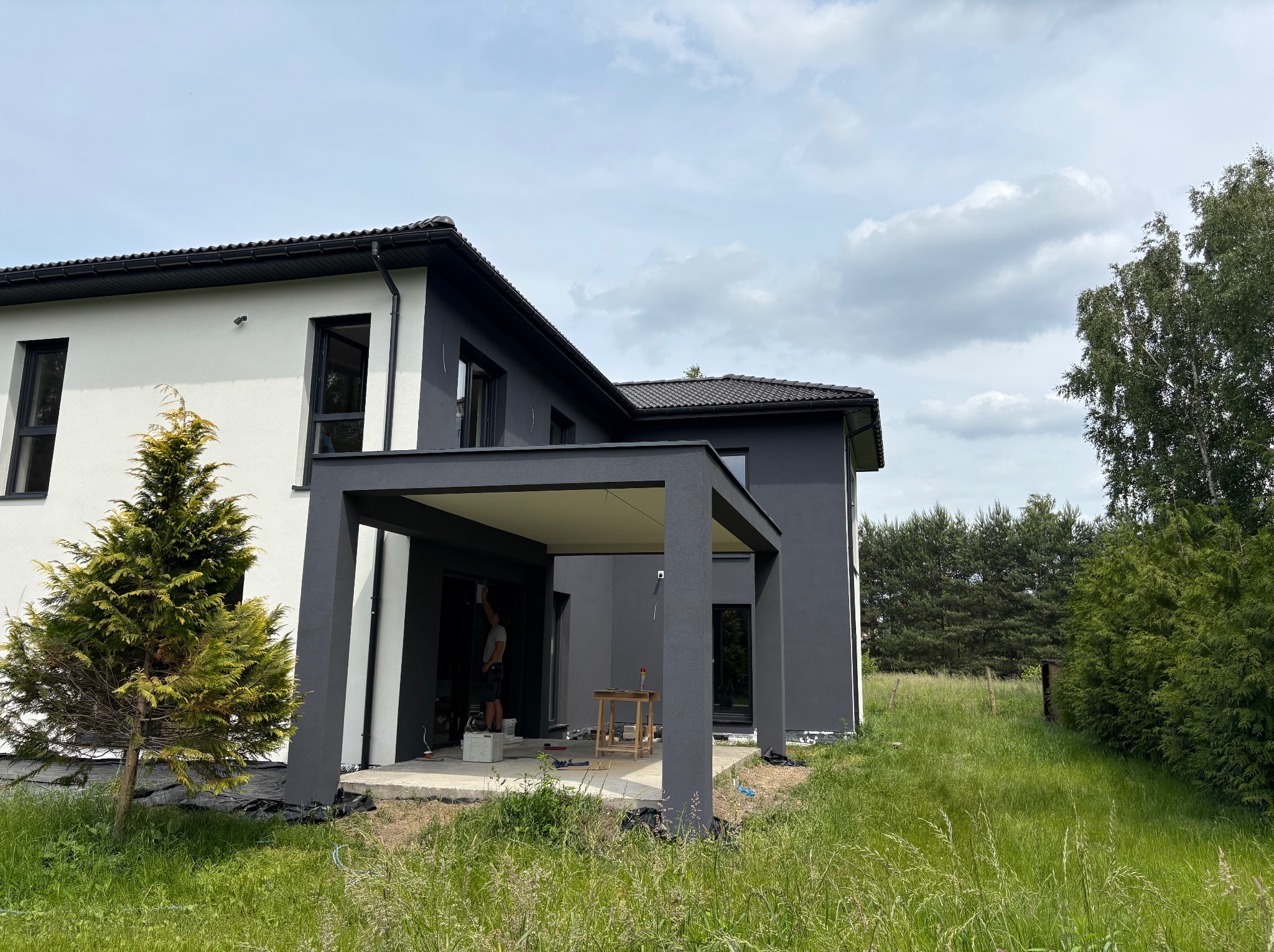 Dwukondygnacyjny dom z płaskim dachem i pergolą, malowany na biało i szaro. Widoczny mężczyzna malujący elewację. Trawa i drzewa w tle.