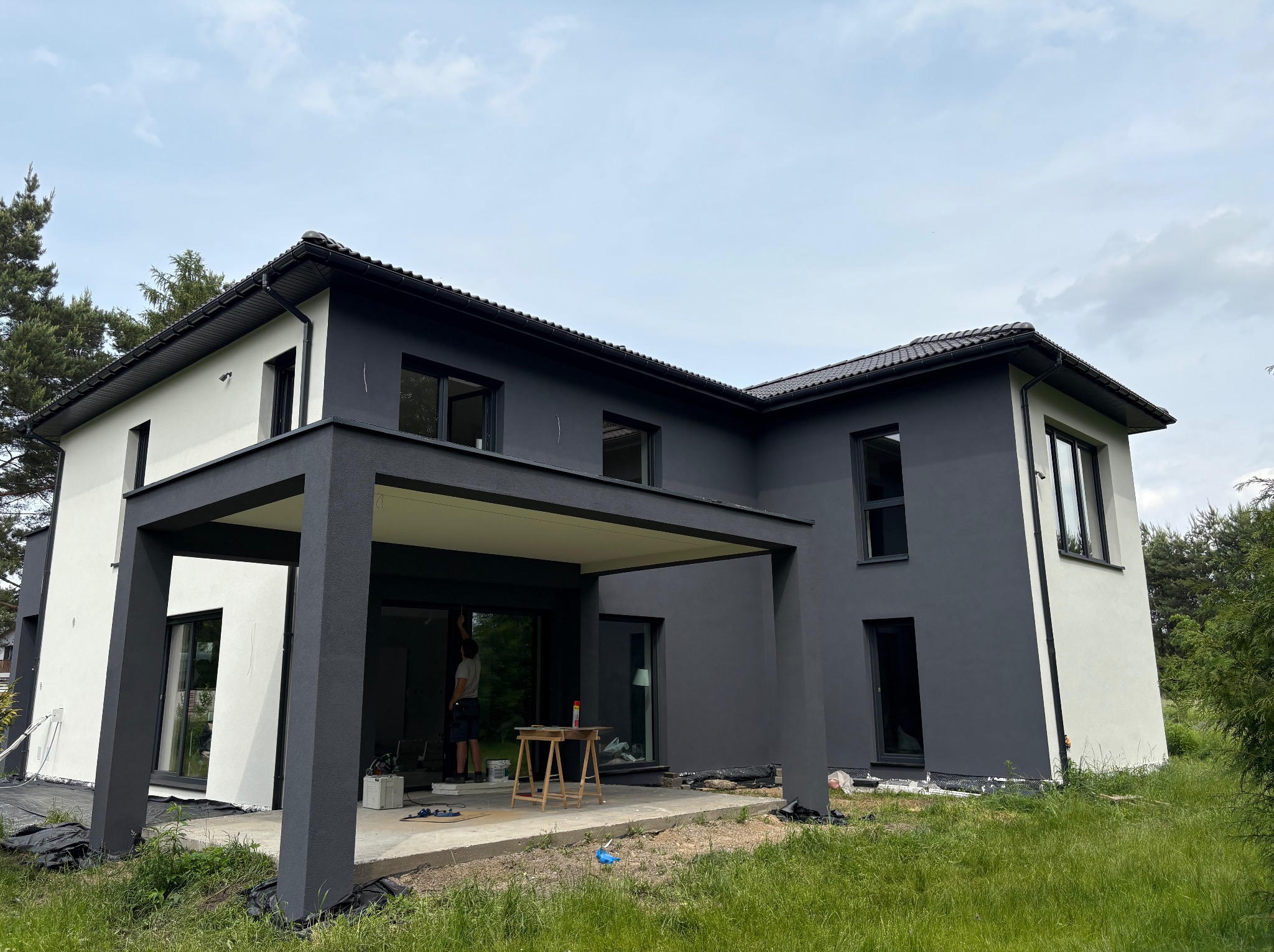 Dwukondygnacyjny dom z płaskim dachem, pomalowany na biało i grafitowo. Widoczny pracownik malujący elewację przy przeszklonym tarasie. Trawa na pierwszym planie.