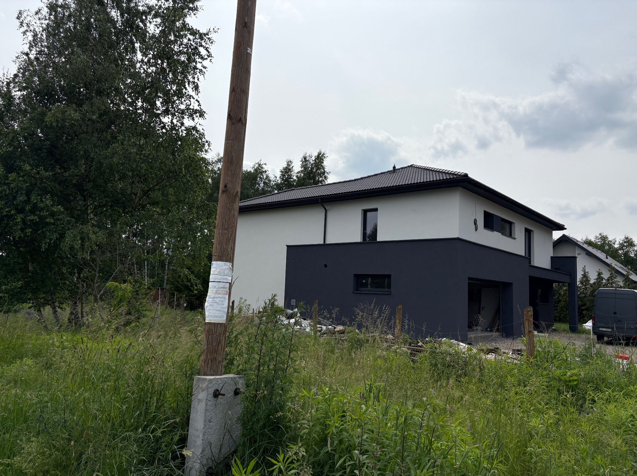 Nowoczesny dom dwukondygnacyjny z płaskim dachem, pomalowany na biało i grafitowo, otoczony wysoką trawą. Widoczny słup energetyczny z ogłoszeniami.