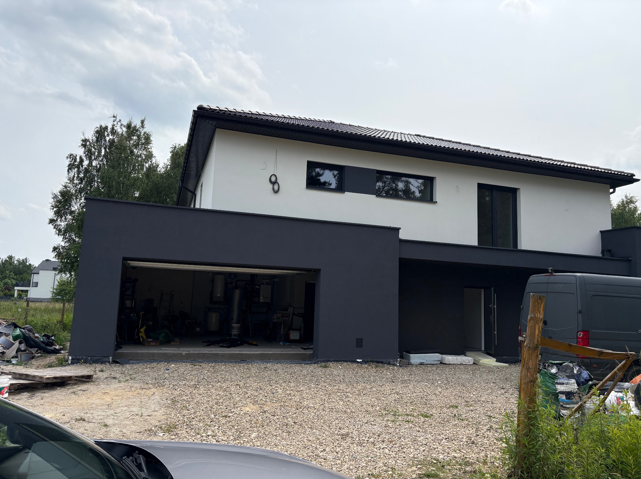 Nowoczesny dom z białą elewacją i grafitowym garażem. Otwarta brama garażowa ukazuje wnętrze. Na zewnątrz gruz i materiały budowlane. Częściowo widoczny samochód.