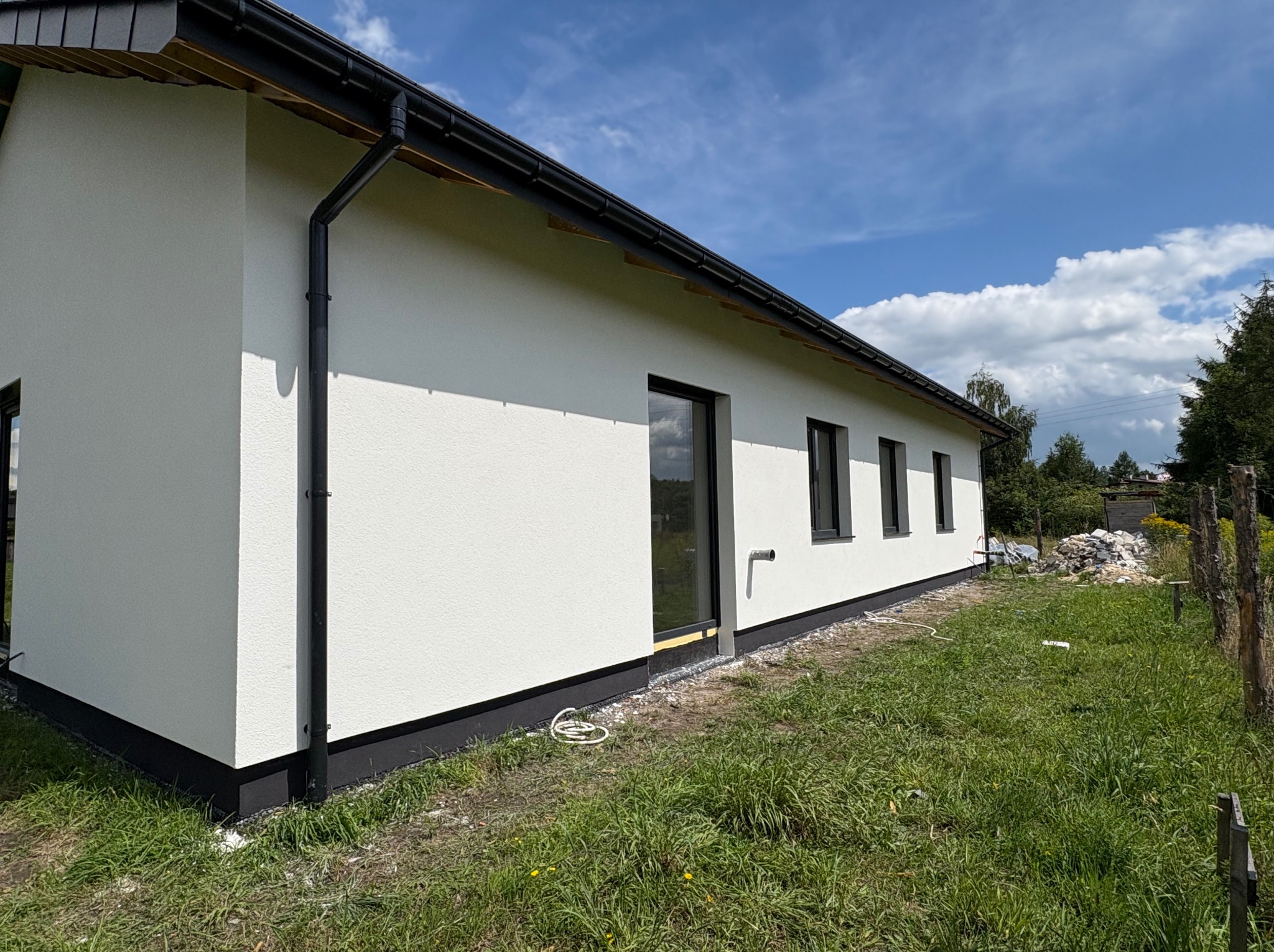 Boczny widok na otynkowany, parterowy dom z czarną stolarką okienną i orynnowaniem. Widoczna faktura tynku i fragment trawnika. Błękitne niebo z chmurami w tle.