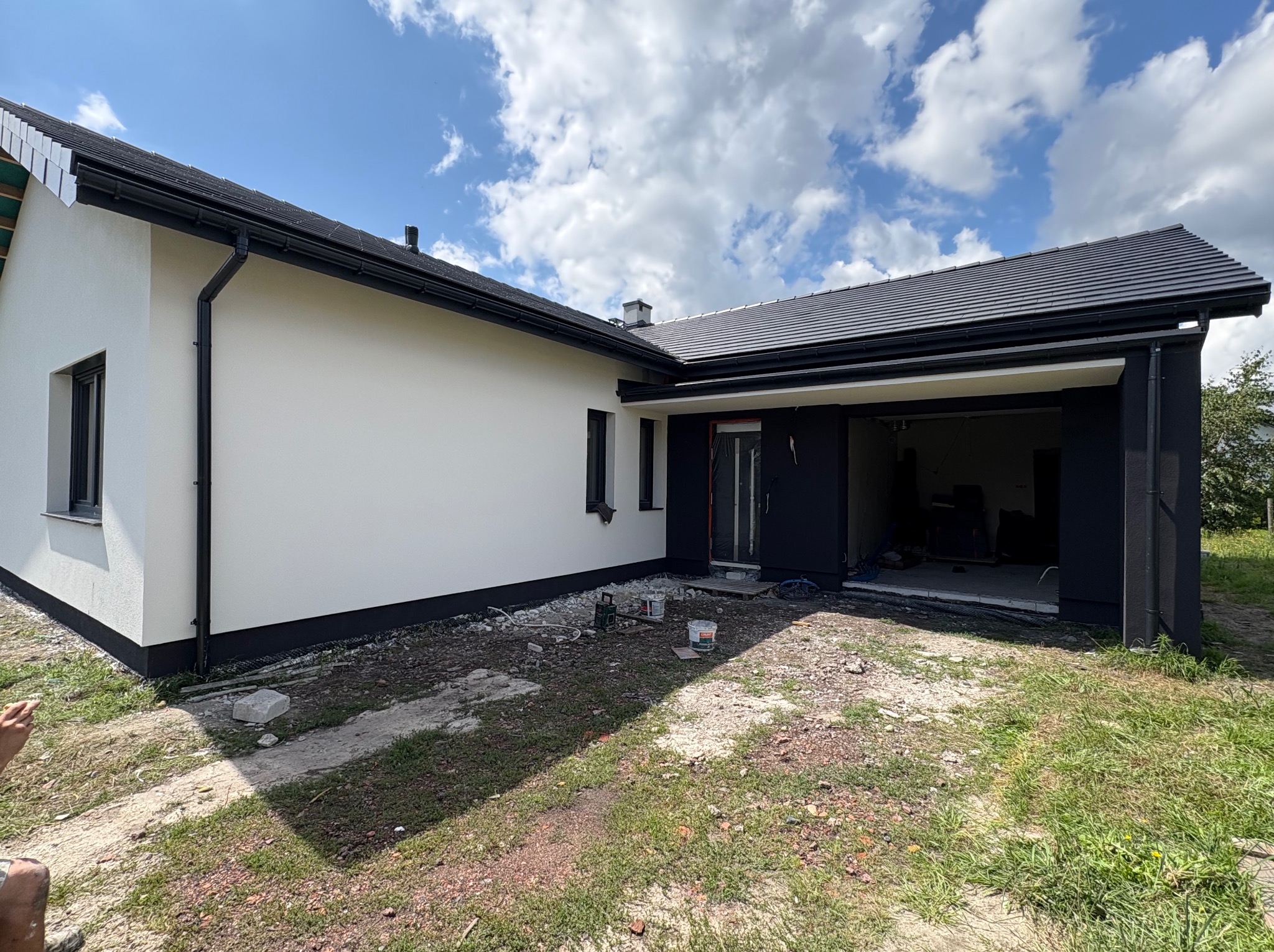 Nowoczesny dom z białą elewacją i czarnymi detalami, widok z boku, teren wokół domu w trakcie prac wykończeniowych, błękitne niebo z chmurami w tle.