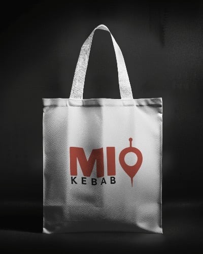 Szara torba z logo 'MIO KEBAB' w kolorze czerwonym, umieszczona na ciemnym tle. Torba z materiału z uchwytami, prezentacja identyfikacji wizualnej.
