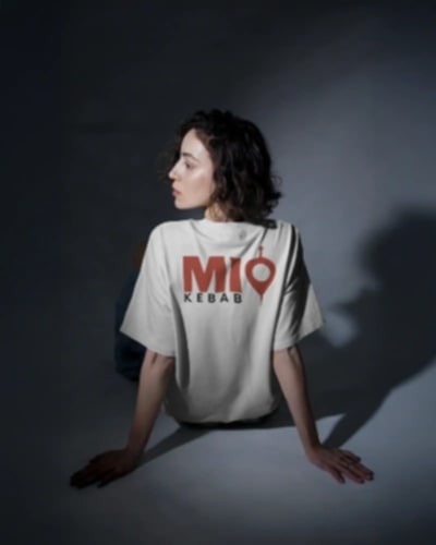 Kobieta siedzi tyłem, ubrana w białą koszulkę z czerwonym logo 'MIO KEBAB' i pinezką lokalizacji. Stonowane oświetlenie, ciemne tło. Minimalistyczny design.