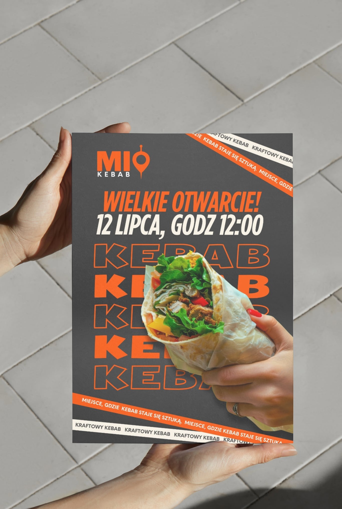 Plakat reklamowy 'Mio Kebab' z informacją o wielkim otwarciu 12 lipca, godz. 12:00, trzymany w dłoniach na tle płytek podłogowych. W centrum grafika kebaba.