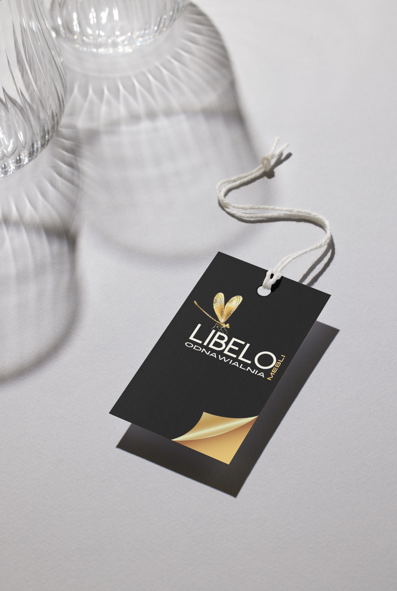 Czarna etykieta z logo 'Libelo Odnawialnia Mebli' ze złotą ważką i podwiniętym rogiem, zawieszona na sznurku, w tle szklane naczynie i cienie.
