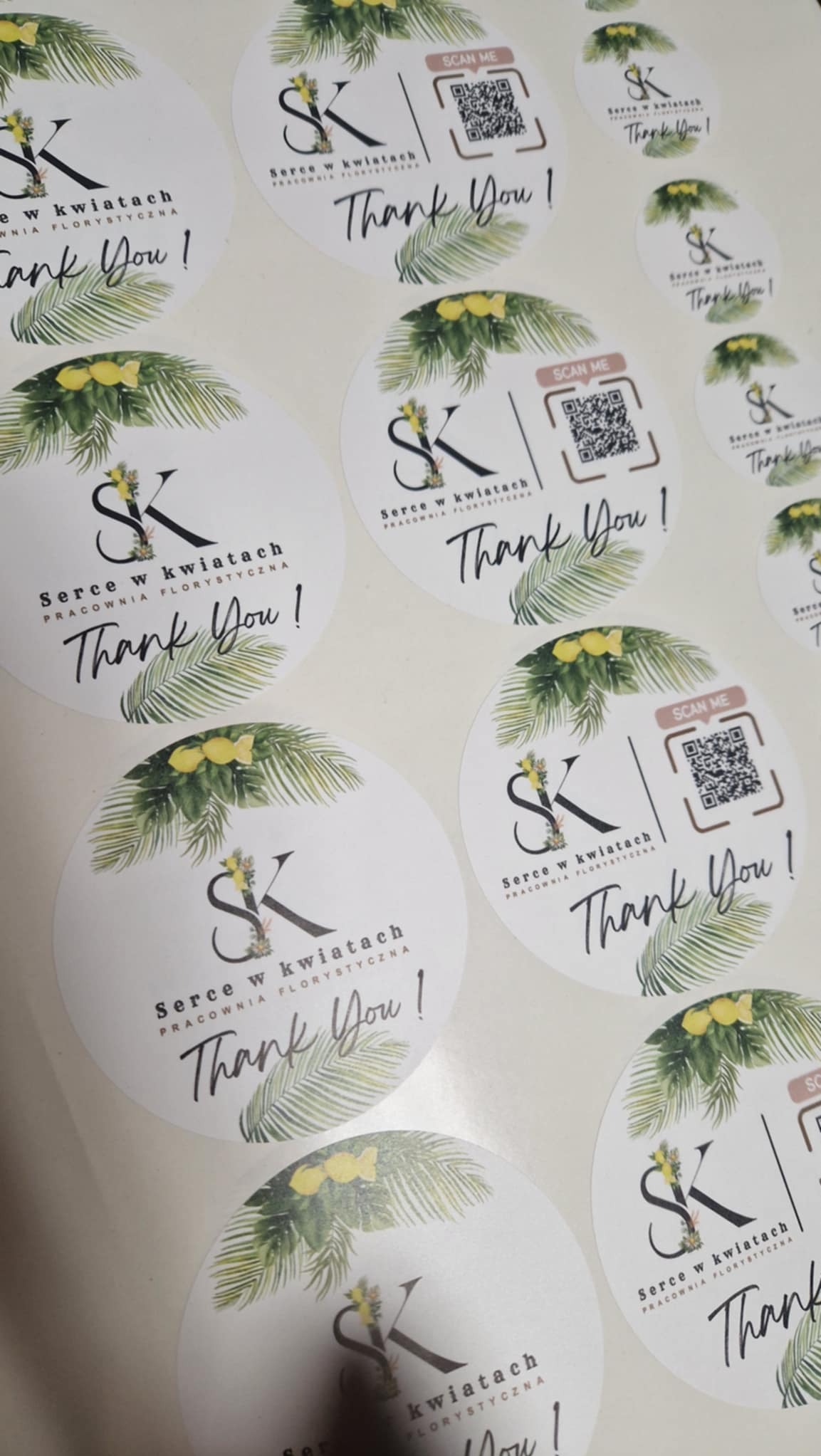 Okrągłe naklejki z logo 'Serce w kwiatach', motywem roślinnym i napisem 'Thank You!' oraz kodem QR. Naklejki na białym tle, w bliskim ujęciu.