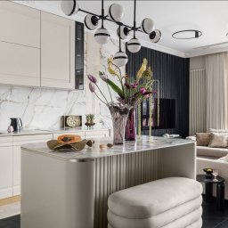 SCM SPÓŁKA Z OGRANICZONĄ ODPOWIEDZIALNOŚCIĄ - Elegancka kuchnia z wyspą i marmurowym blatem, kremowe szafki, czarne baterie, dekoracyjny wazon z kwiatami, widok na salon z sofą i oknem.