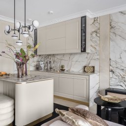 SCM SPÓŁKA Z OGRANICZONĄ ODPOWIEDZIALNOŚCIĄ - Elegancka kuchnia w beżach z marmurowym blatem i ścianą. Wyspa kuchenna z kwiatami, nowoczesne oświetlenie, czarne akcenty i dekoracje tworzą luksusowy klimat.