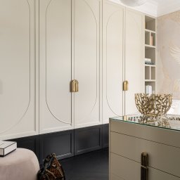 SCM SPÓŁKA Z OGRANICZONĄ ODPOWIEDZIALNOŚCIĄ - Beżowa szafa wnękowa z czarnym dołem i złotymi uchwytami, uzupełniona pufą i komodą z dekoracyjną misą, na tle parkietu w jodełkę i ozdobnej tapety.