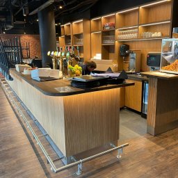 SCM SPÓŁKA Z OGRANICZONĄ ODPOWIEDZIALNOŚCIĄ - Nowoczesny bar z drewnianym blatem i podświetlanym frontem. W tle półki z oświetleniem, szklanki i ekspres do kawy. Widoczny fragment ceglanej ściany i krzesła barowe.