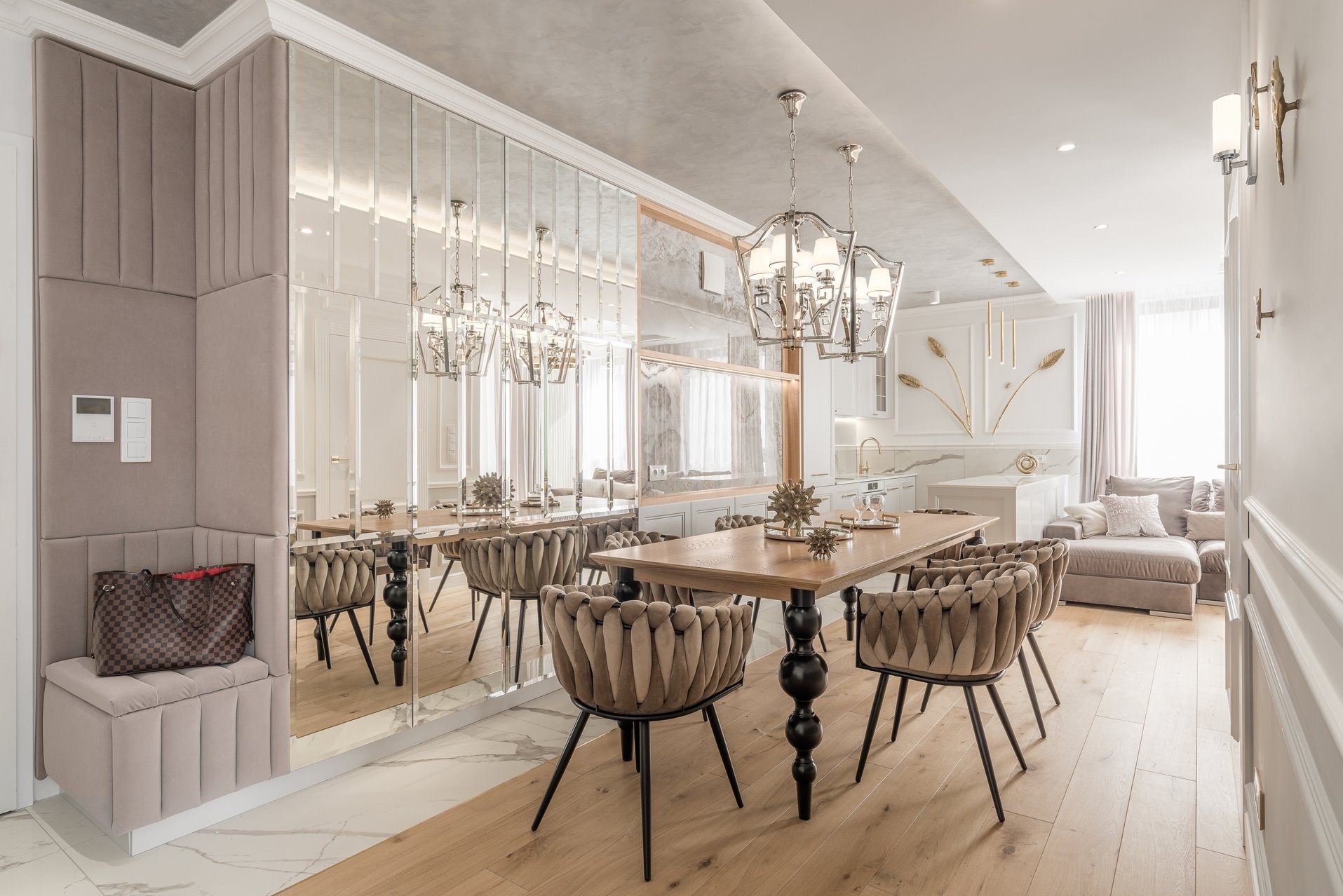 Elegancka jadalnia z lustrzaną ścianą, drewnianym stołem i tapicerowanymi krzesłami. W tle widoczna kuchnia i salon w jasnych barwach. Całość w stylu glamour.