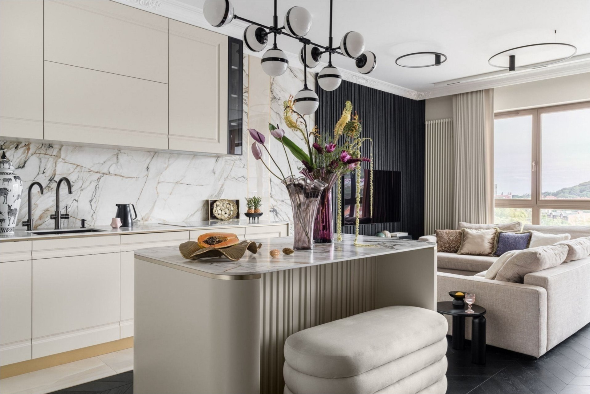 Elegancka kuchnia z wyspą i marmurowym blatem, kremowe szafki, czarne baterie, dekoracyjny wazon z kwiatami, widok na salon z sofą i oknem.