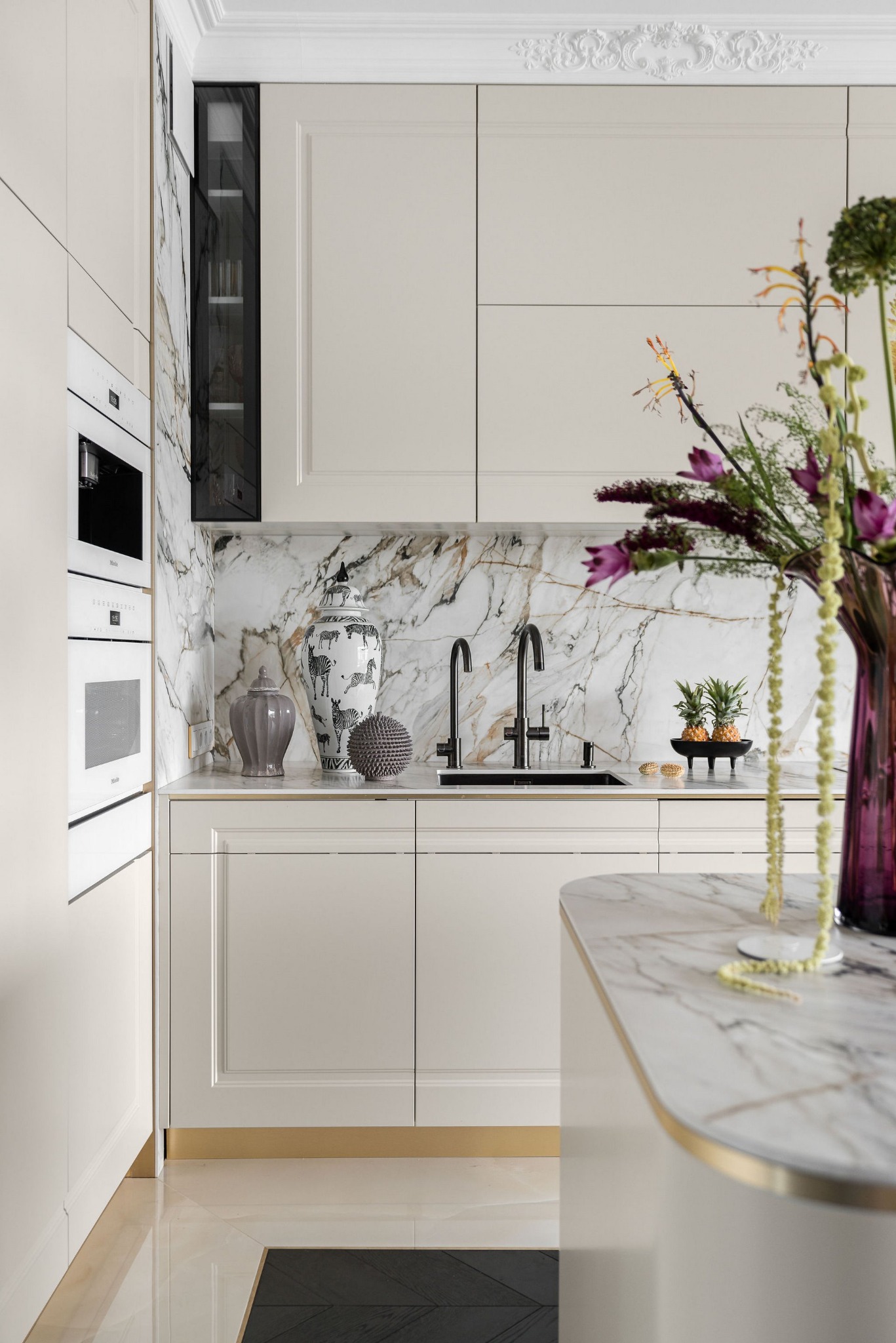 Elegancka kuchnia w odcieniach beżu z marmurowym blatem i backsplashiem. Uwagę zwracają dekoracyjne wazony i świeże kwiaty, dodające wnętrzu luksusowego charakteru.