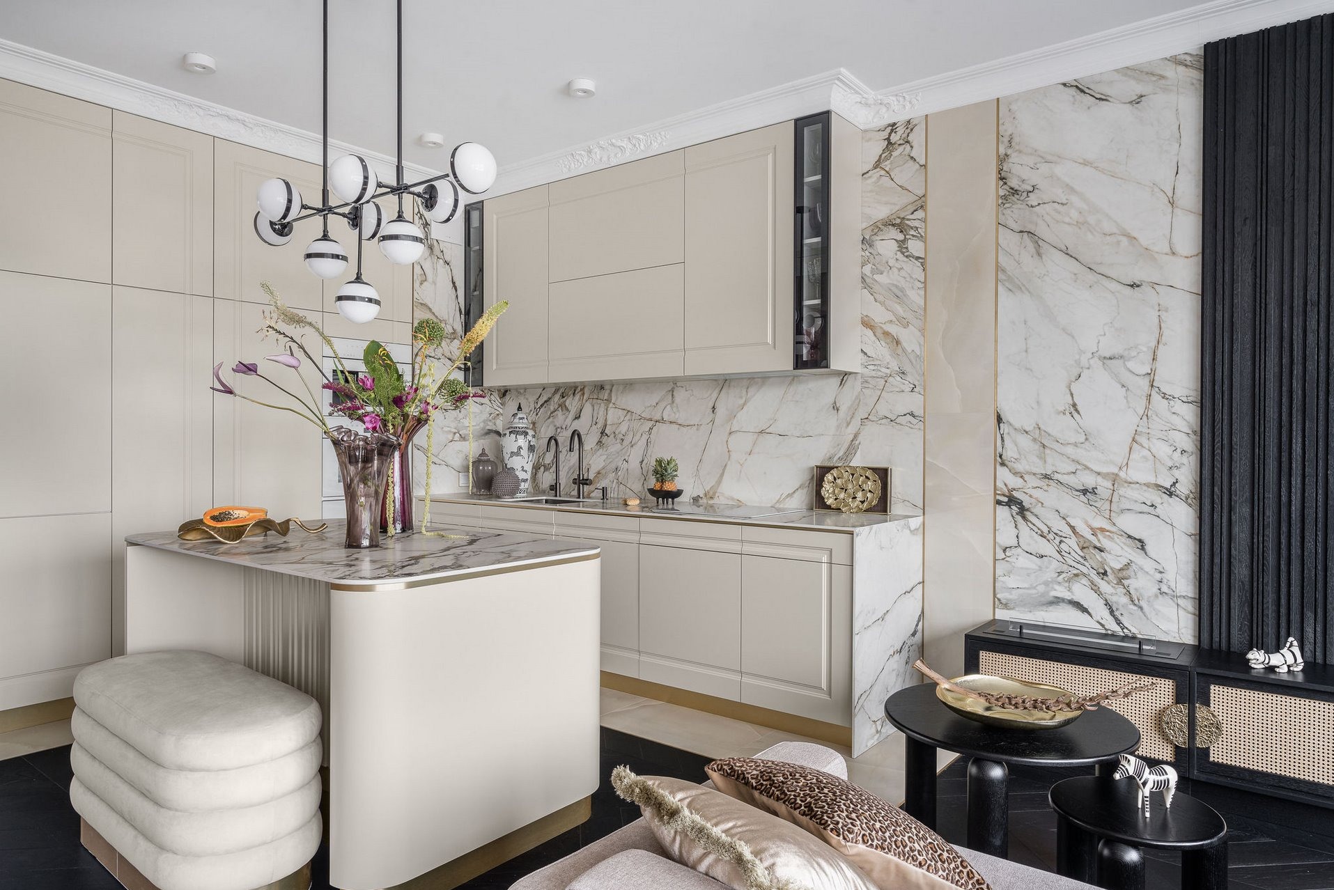 Elegancka kuchnia w beżach z marmurowym blatem i ścianą. Wyspa kuchenna z kwiatami, nowoczesne oświetlenie, czarne akcenty i dekoracje tworzą luksusowy klimat.