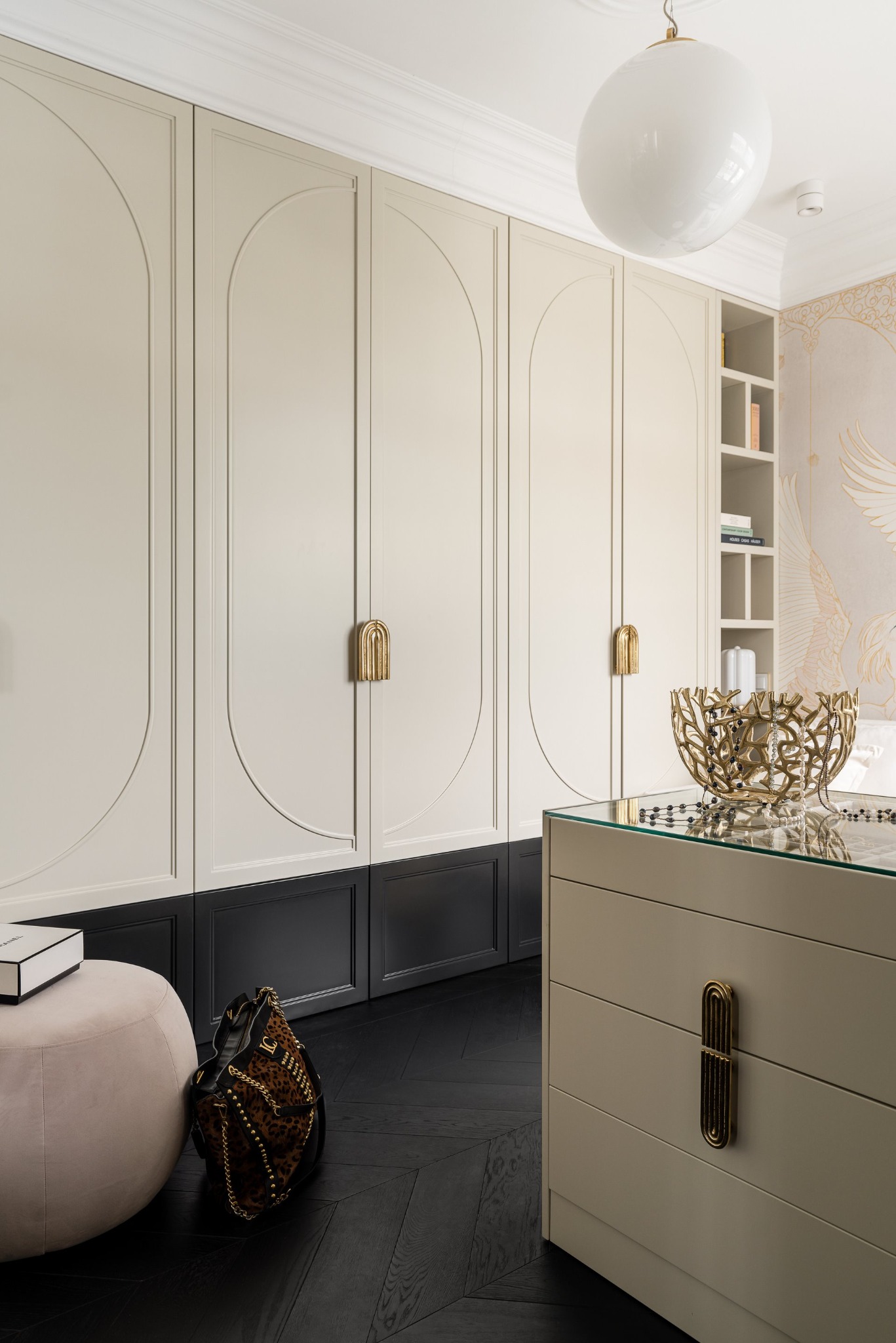 Beżowa szafa wnękowa z czarnym dołem i złotymi uchwytami, uzupełniona pufą i komodą z dekoracyjną misą, na tle parkietu w jodełkę i ozdobnej tapety.