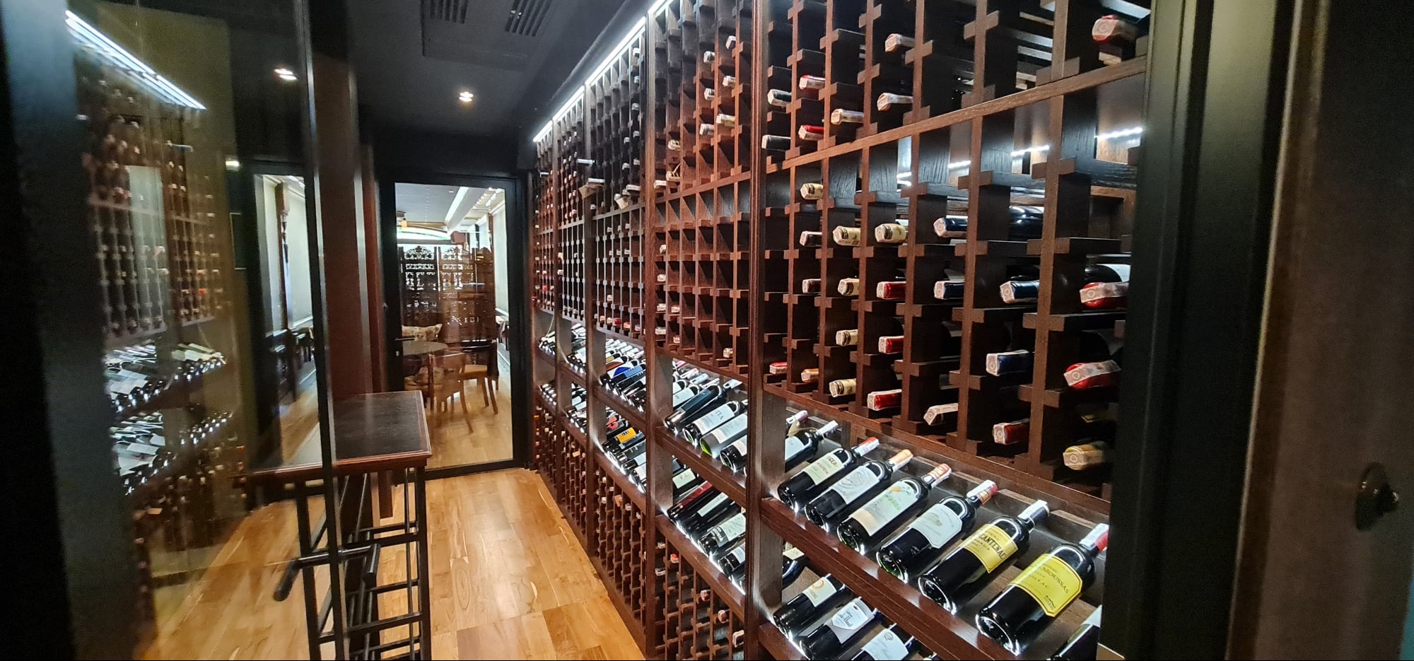 Drewniana zabudowa winiarni z licznymi półkami na butelki wina, widok z perspektywy korytarza. W tle widoczne pomieszczenie z krzesłami i stołem.