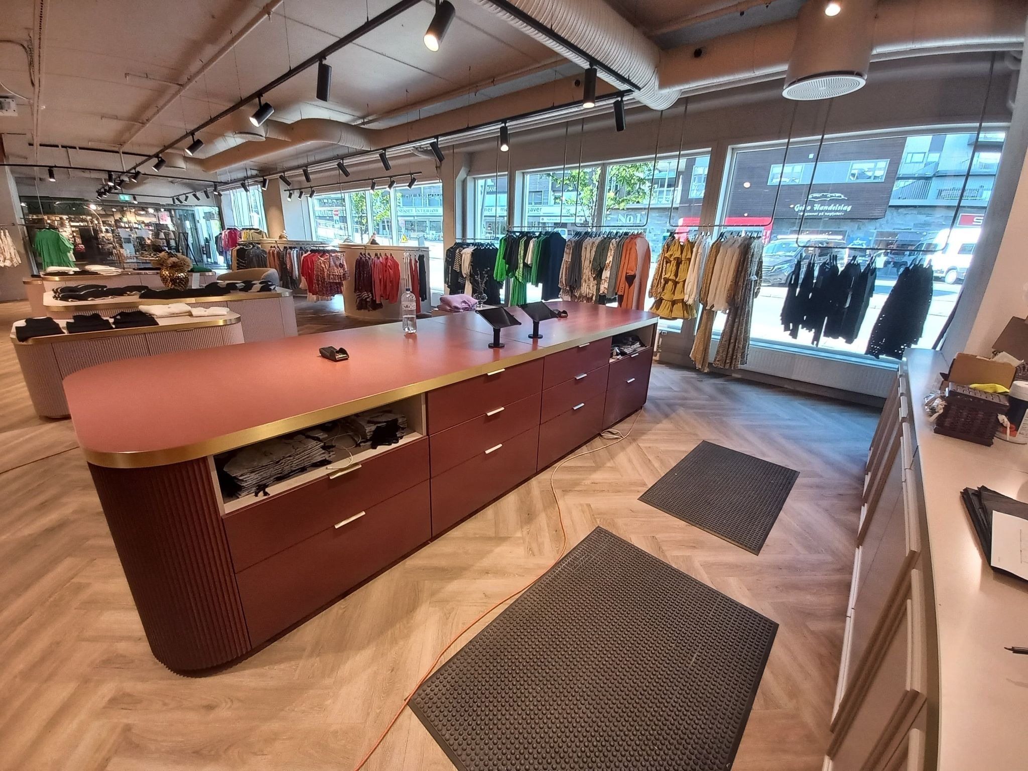 Wnętrze sklepu odzieżowego z kasą/ladą w kolorze bordo z złotymi akcentami. W tle wieszaki z ubraniami i duże okna. Podłoga w jasnym odcieniu drewna.