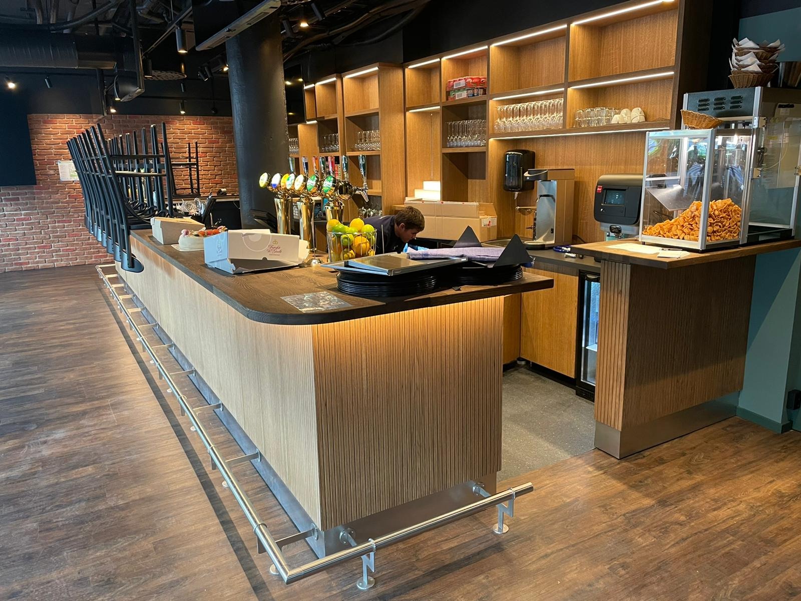 Nowoczesny bar z drewnianym blatem i podświetlanym frontem. W tle półki z oświetleniem, szklanki i ekspres do kawy. Widoczny fragment ceglanej ściany i krzesła barowe.