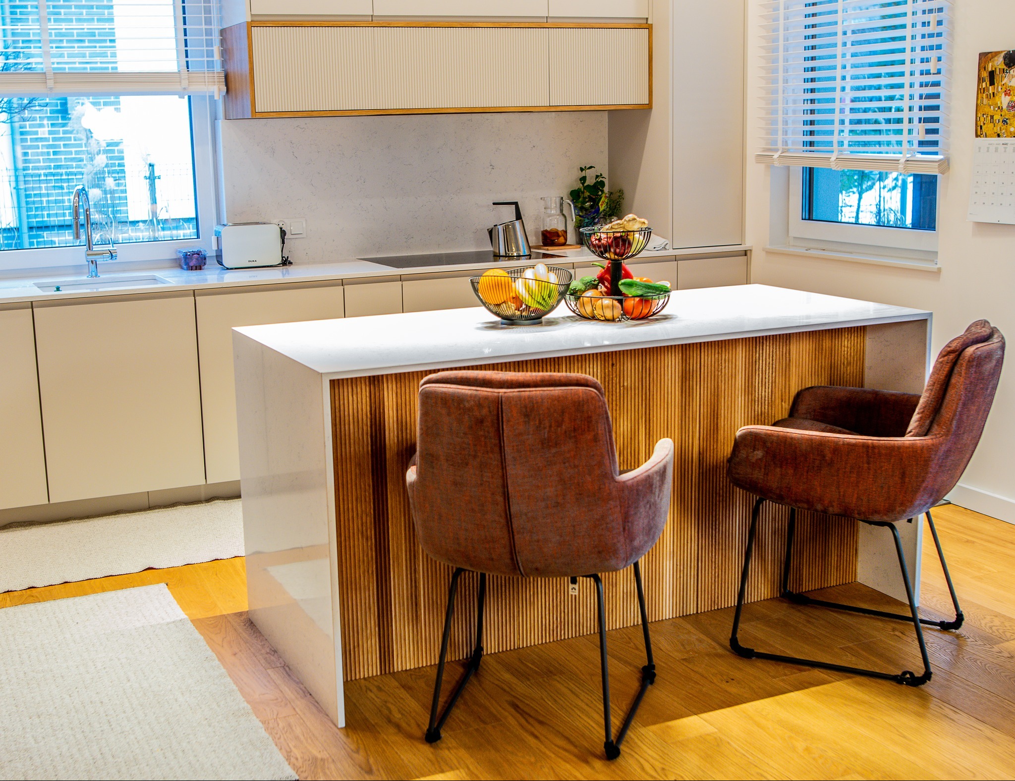 Nowoczesna kuchnia z wyspą: biały blat, drewniane panele, krzesła barowe w odcieniach brązu. Owoce w koszykach na blacie. Jasne, naturalne światło.