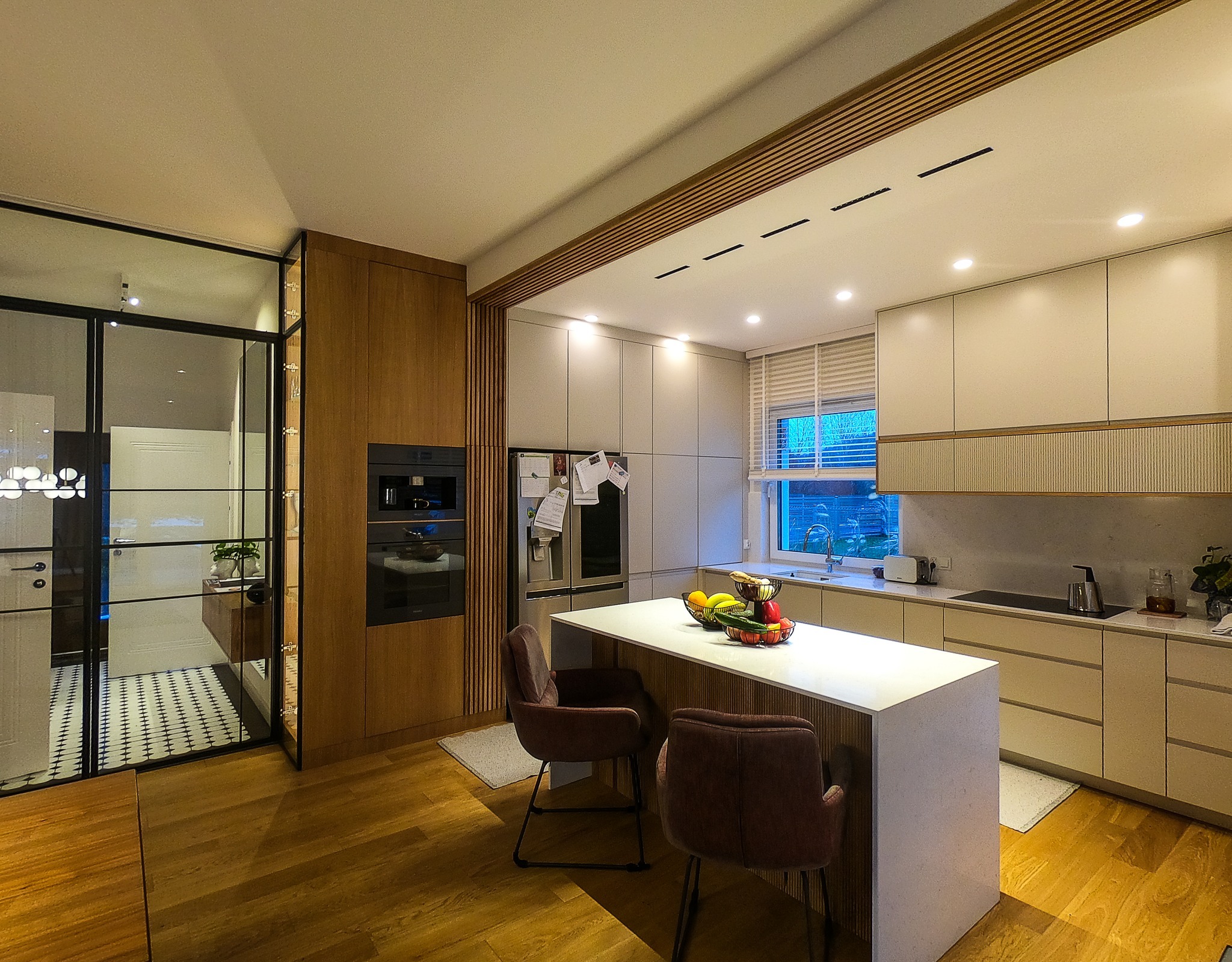 Nowoczesna kuchnia z wyspą i drewnianymi akcentami. Widoczne przeszklone drzwi i oświetlenie punktowe. Na wyspie kosze z owocami.