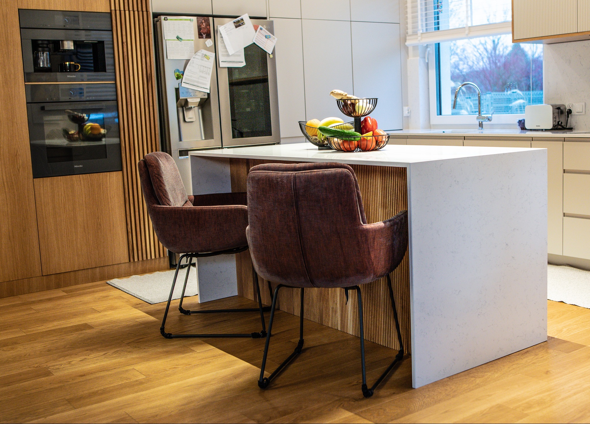 Nowoczesna kuchnia z wyspą i dwoma krzesłami w odcieniach brązu. Na blacie wyspy koszyk z owocami. W tle zabudowa kuchenna z drewnianymi akcentami i sprzętem AGD.