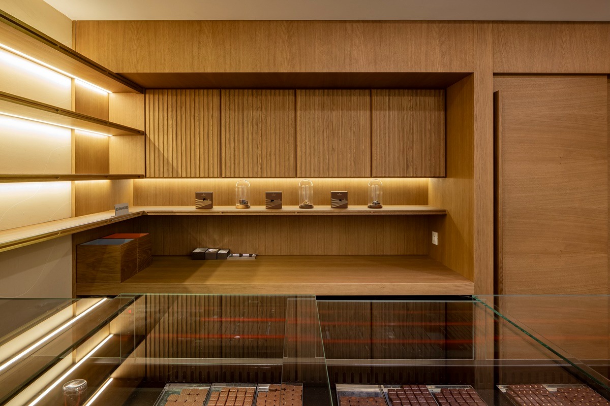 Drewniane meble na wymiar w cukierni: szafki z ryflowanymi frontami, podświetlane półki z ekspozycją słodyczy za szklaną witryną. Ciepłe oświetlenie.