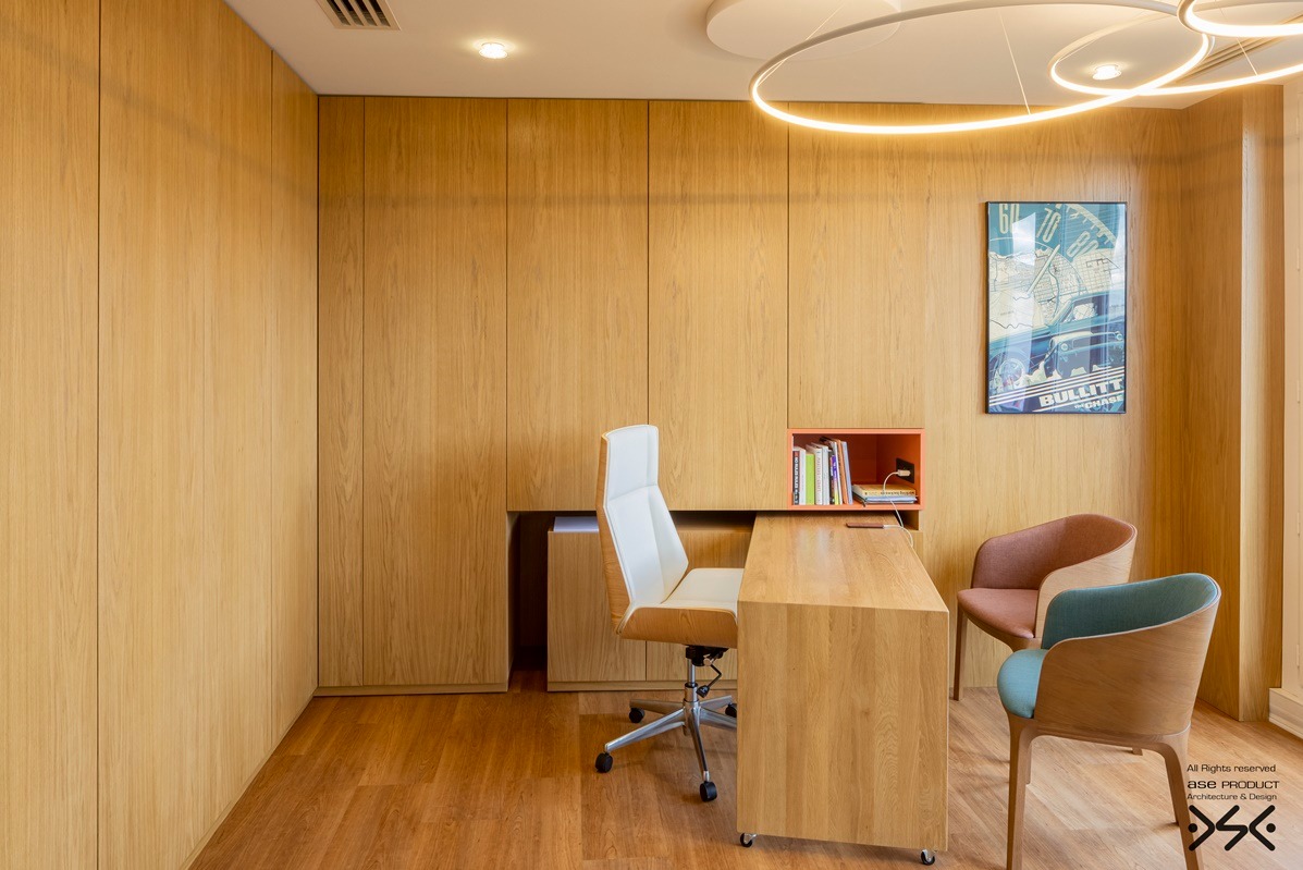 Jasne biuro z drewnianą zabudową: biurko, krzesła, szafki wnękowe. Nad biurkiem półka z książkami i plakat filmowy. Nowoczesne oświetlenie sufitowe.