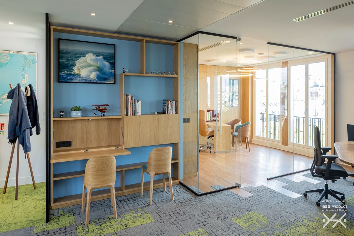 Nowoczesne biuro z niebieską ścianą, drewnianymi meblami i szklaną ścianą działową. Na ścianie obraz z motywem morskim. Widoczne krzesła i dywan w geometryczne wzory.