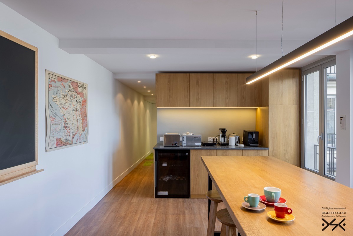 Jasna kuchnia z drewnianymi szafkami i blatem, oświetlona listwą LED. Na stole filiżanki, w tle mapa Francji i tablica. Nowoczesny design z elementami vintage.