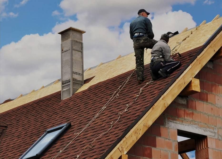 Dekarze na dachu z brązową dachówką, zabezpieczeni linami, pracują przy kominie. Widoczny fragment muru i okno dachowe.
