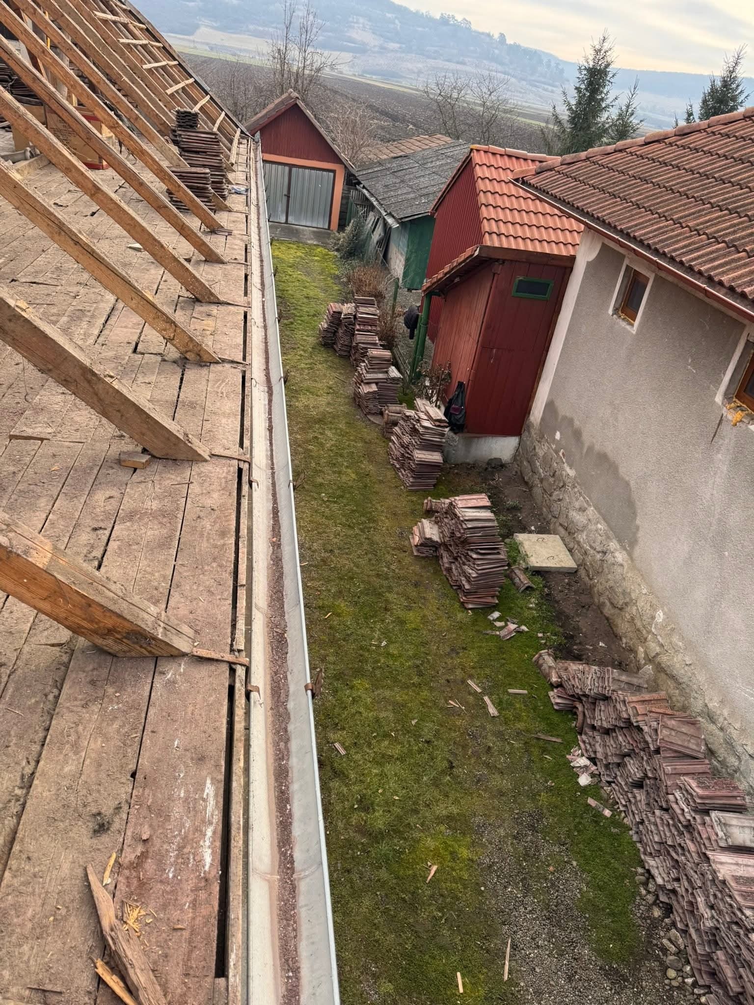 Rynna zamontowana na dachu z drewnianą konstrukcją, obok sterty dachówek i budynki w tle. Widok z góry na rynnę i przygotowania do prac dekarskich.