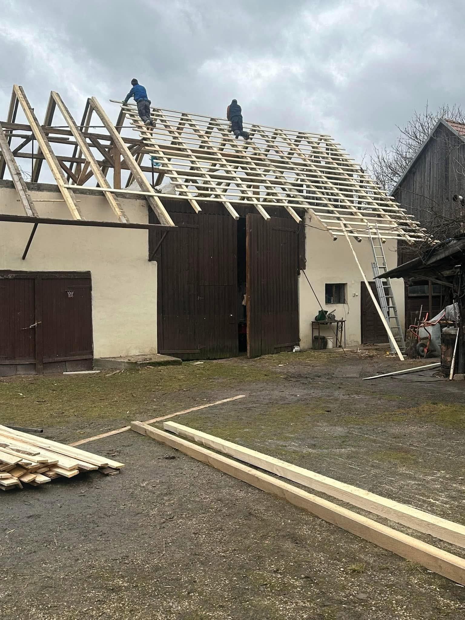 Dwóch dekarzy na drewnianej konstrukcji dachu budynku gospodarczego. Widoczne deski, drabina i narzędzia. Pochmurne niebo w tle. Prace dekarskie w toku.