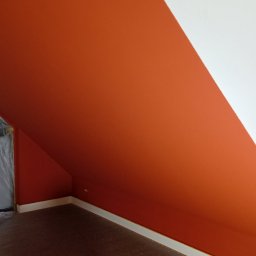 PRZEDSIĘBIORSTWO USŁUGOWO-BUDOWLANE KAMIL KUBICKI - Pomarańczowo-białe malowanie skośnej ściany poddasza, okno zabezpieczone folią malarską, widoczne taśmy malarskie, podłoga zabezpieczona.