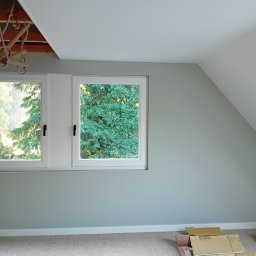 PRZEDSIĘBIORSTWO USŁUGOWO-BUDOWLANE KAMIL KUBICKI - Pokój na poddaszu po malowaniu ścian na jasny, pastelowy kolor. Widoczne okna z widokiem na zieleń, drewniane elementy konstrukcyjne sufitu i kartony na podłodze.
