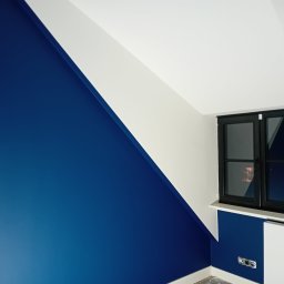 PRZEDSIĘBIORSTWO USŁUGOWO-BUDOWLANE KAMIL KUBICKI - Ściana w odcieniach bieli i intensywnego granatu w pokoju na poddaszu z oknem w czarnej ramie. Widoczne gniazdka elektryczne i fragment grzejnika.